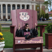 Lisa K in grad cap and gown sitting in large oversized branded Goldy Gopher chair on Northrop Mall