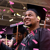 a man celebrates his graduation in a cap and gown with pink confetti falling around him