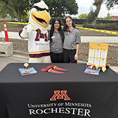 two umr students and a mascot stand behind a U of M Rochester table