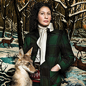 a woman wearing a suitcoat and scarf with a painted wooded background and a stuff coyote in front of her