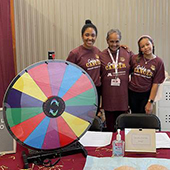 Aleta S and two coworkers behind a presentation table that includes a wheel of fortune like spinning wheel