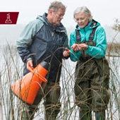 Two senior citizens in waders stand in shallow water collecting samples
