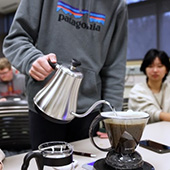 a person pouring coffee from a silver pot as a student looks on