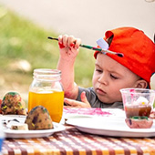 a toddler in a baseball cap holds a paintbrush at a craft table
