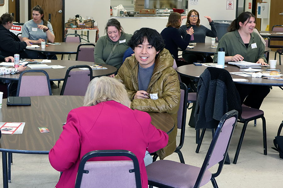 A man sits and talks with an elderly woman in the foreground and a group of women seated at tables and talking in the background.