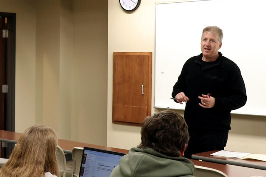 Mark Collier stands at the front of a classroom teaching to students.