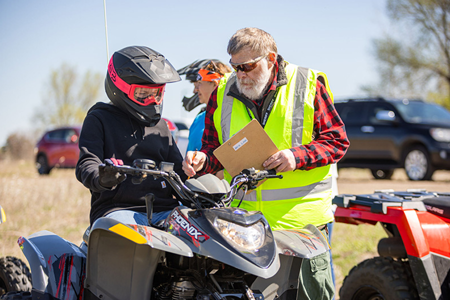 A young rider in a helmet receives instruction from an older man in a yellow vest in a grassy outdoor setting.