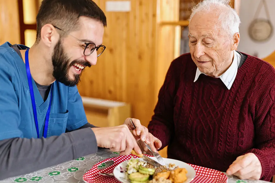 Younger male care worker sitting at a table with older man helping him eat a meal.