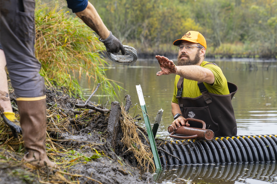 Afton Clarke-Sather reaches for coiled wire from another person's hand while standing in a lake next to a large black tube.