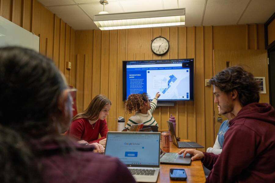 Research group seated around a table with one student pointing towards a presentation screen.