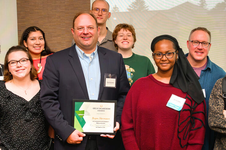 ryan Hermann holds an award plaque in the middle of students and staff. 